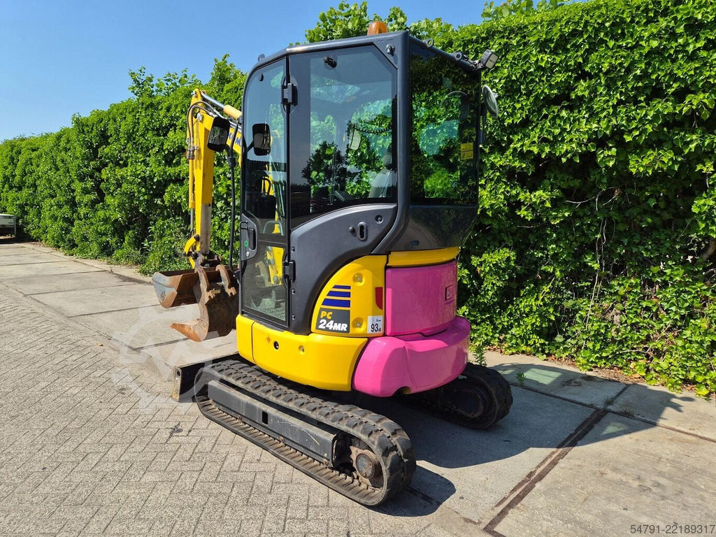 Mini-pelle sur chenilles Ma boutique Komatsu PC24MR-5 jaune et rose avec cabine vitrée sur un chantier extérieur